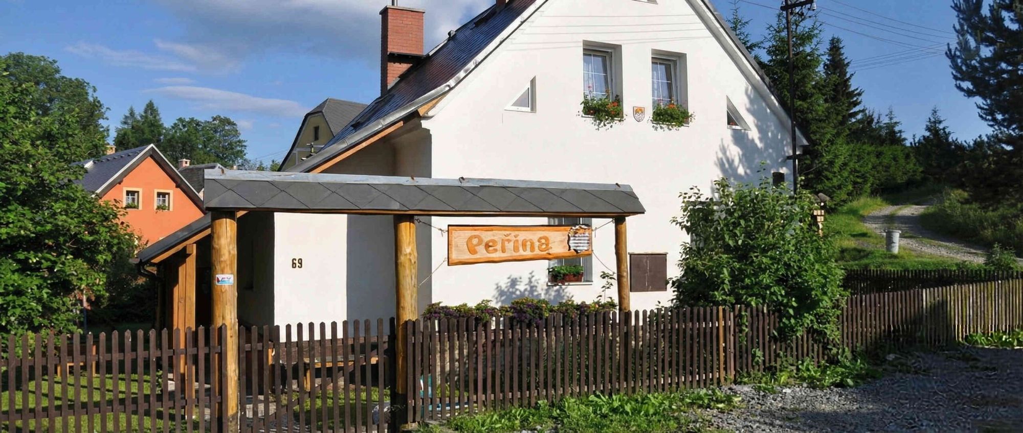 Bílá chalupa Peřina s dřevěnou cedulí nad vstupem, kolem dokola dřevěný plot a zeleň
